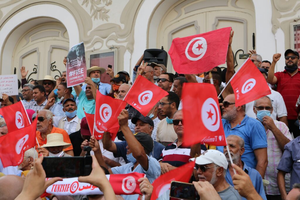Impression de drapeaux tunisiens – Fabrication en papier et tissu pour événements et institutions Foule brandissant des drapeaux tunisiens rouges et blancs lors d’un rassemblement, illustrant la qualité et la visibilité des impressions de drapeaux fabriqués en Tunisie.